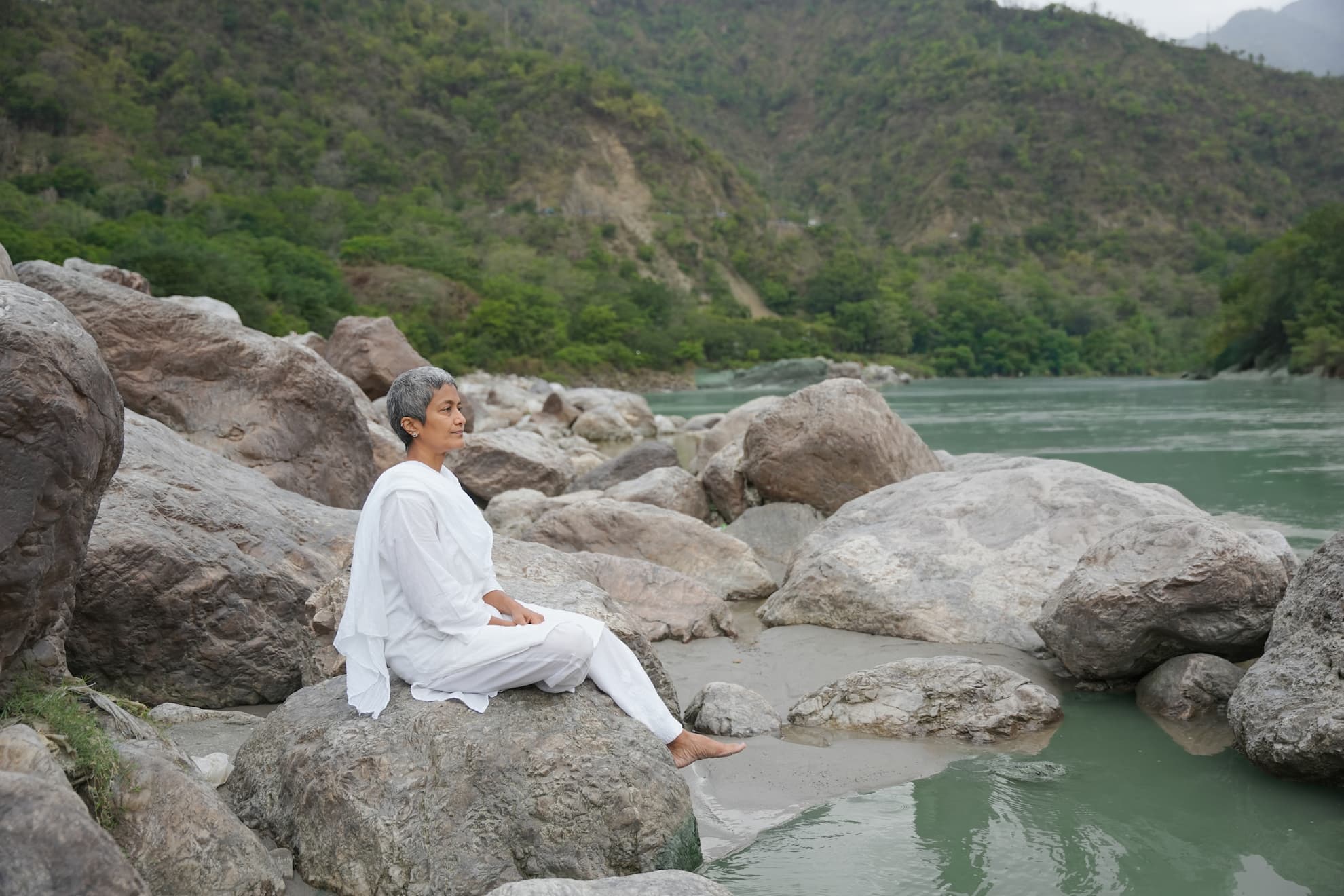 Sashi Kumar, founder of Rupin Travels, seated cross-legged on the ghats of Rishikesh in a white kurta, Laxman Jhula bridge and the Ganges visible behind her