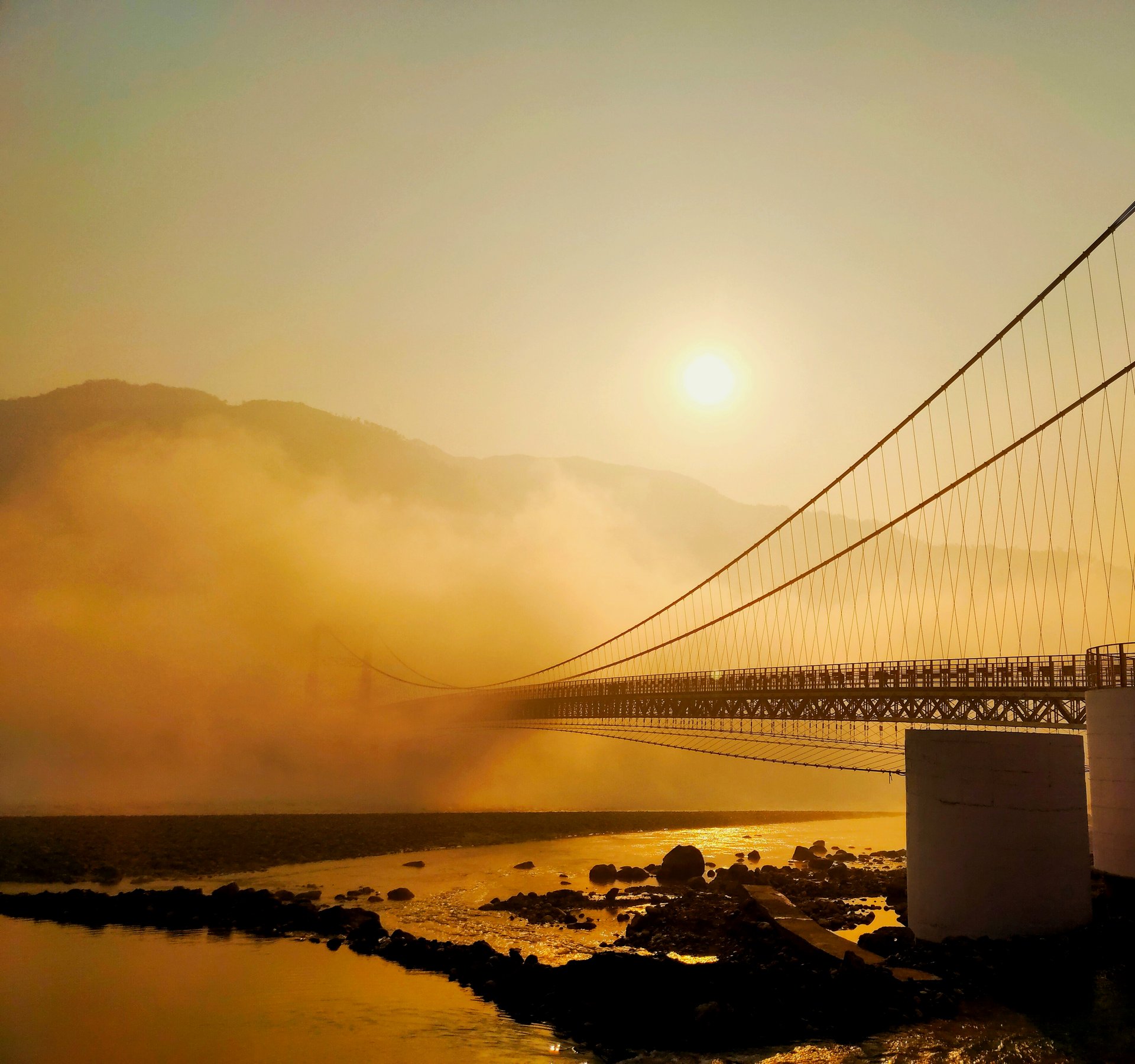 Sunrise over the Ganges at Rishikesh — golden mist rising off the river, Ram Jhula suspension bridge silhouetted against the morning light, Himalayan hills behind
