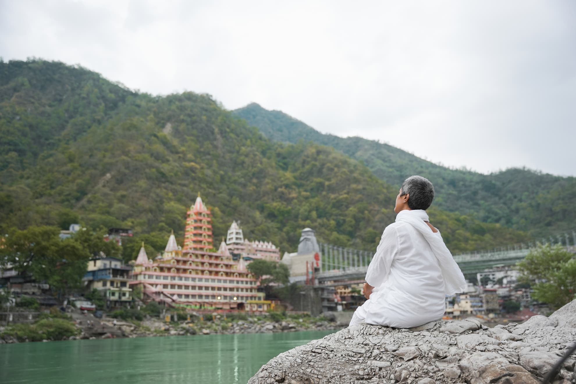 Woman in white kurta seated on the ghats of Rishikesh looking across the Ganges toward Trayambakeshwar temple and Ram Jhula bridge in morning mist