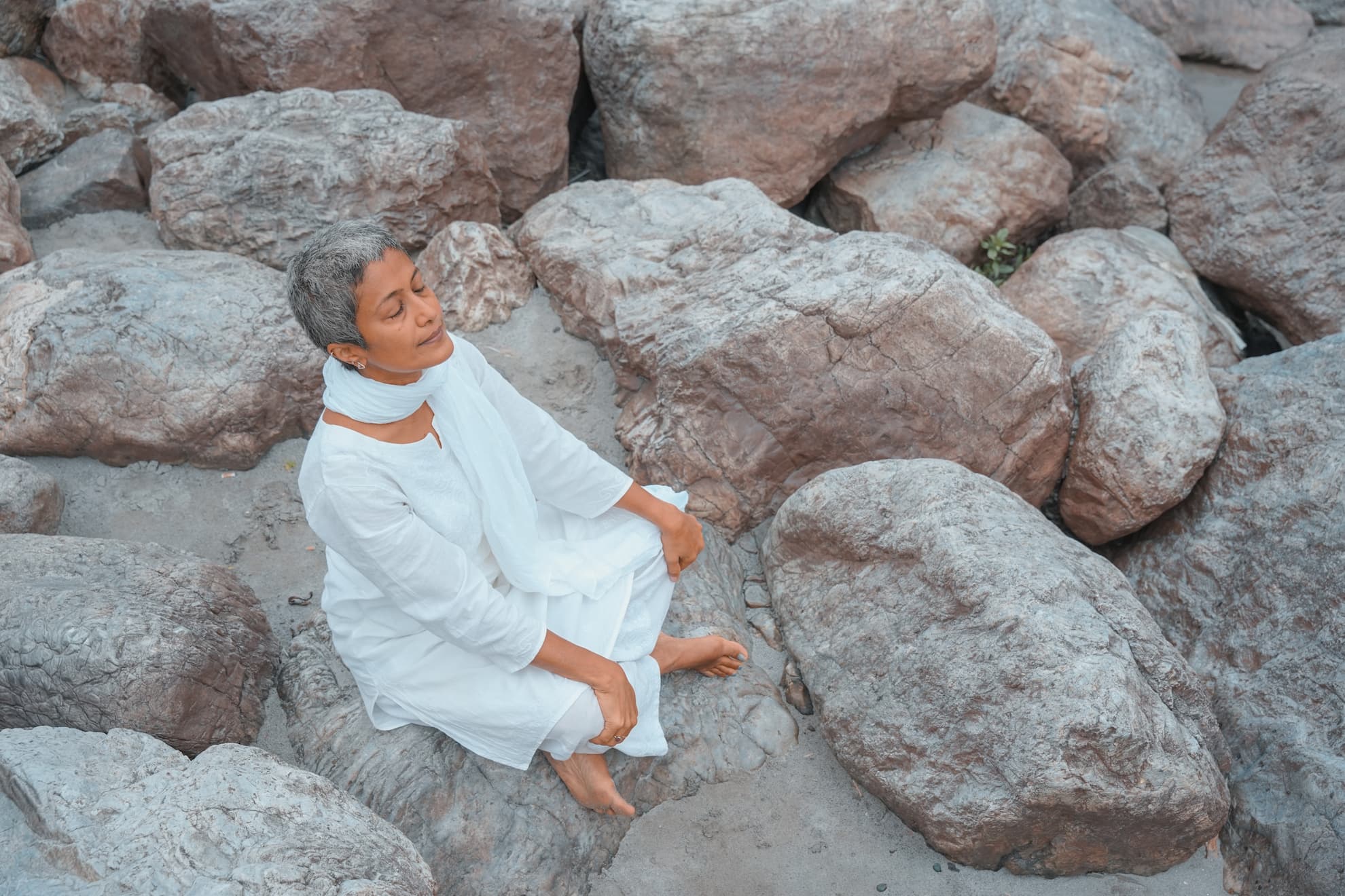 Sashi Kumar, founder of Rupin Travels, seated cross-legged on the ghats of Rishikesh in a white kurta, Laxman Jhula bridge and the Ganges visible behind her