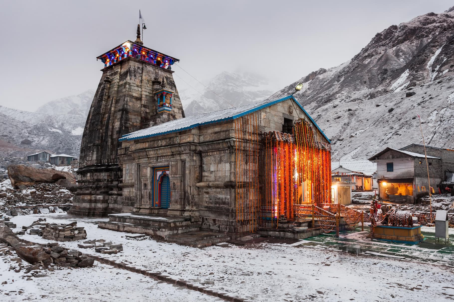 Kedarnath temple covered in snow with marigold garland decorations glowing against the glacial Himalayan peaks at dusk